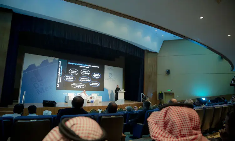 يسلط الضوء على شريحة على شرائح بيانية أثناء تواجده على منصة ملتقى طب الأسنان، وذلك في قاعة محاضرات حديثة ومضيئة في القصيم، بينما يتابع الحضور باهتمام، ومن بينهم عدد من المشاركين اضهر الغترة أو الشماغ السعودي مع المقال والبشت السعودي.