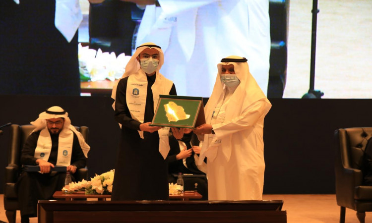 رجلان يرتديان الغترة أو الشماغ السعودي مع العقال والبشت السعودي على خشبة المسرح في جامعة القصيم، وهما يضعان الكمامات الطبية التزاماً بإجراءات الاحترازية. ويقوم بتسليم الآخر درعاً تكريمياً مُؤطراً لمنطقة المملكة العربية السعودية، وذلك بحضور عدد من الحضور المشاركين في برنامج "السفراء الوسطي"، أثناء توقيت شاشة عرض كبيرة في منصة البناء.