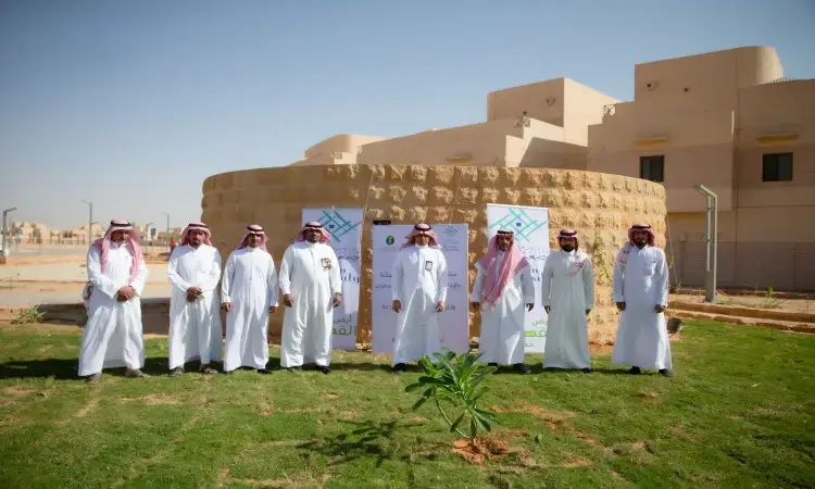 A miami group of men in ghatta, shamma, aqal and Saudi winter shirts on the green lawns in front of the Hajari Circle Building, surrounded by banners highlighting Qassim University's efforts to preserve the environment, while residential buildings and clear skies frame the scene.