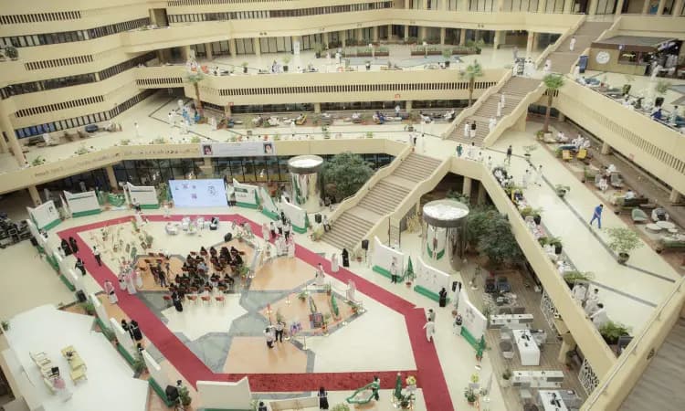 The expansive atrium is home to the university's exhibition, with the event area featuring informational displays, seating for guests, musical entertainment, and a vendor's corner dedicated to the seventh cream. The presence of a ghatta or shemagh with an aqal, along with members of the Saudi winter, including a follow-up with the majesty of the target. Stairs, plants, and streams are scattered throughout this open, modern architectural statement, reflecting the welcoming and uplifting spirit of Qassim University.