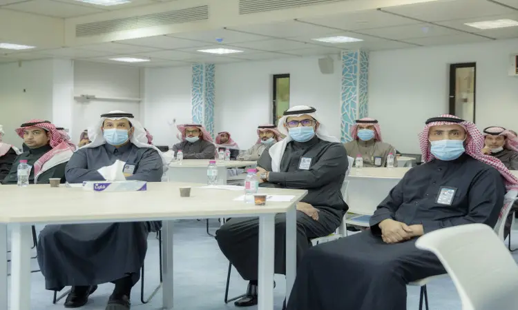اجتمع عدد من الرجال مرتدين الغترة أو الشماغ والعقال، بالإضافة إلى البشت السعودي، وهم ملتزمون بارتداء الكمامات، للمشاركة في دورة عمل في كلية الاقتصاد والإدارة في القصيم. وقد يتم عقد جلسة في قاعة الاجتماع الحديثة لتجهيز الاجتماعات احتوت على طاقم المياه والمناديل الورقية الورقية على الطاولات، وقادرون جميعاً باهتمام نحو القاعة.