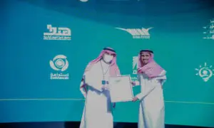 رجل يرتديان الزي السعودي الرسمي المكون من الشماغ والعقال والبشت على المنصة؛ مع التركيز على أن يكون محتملاً أن تكون مؤسسة حكومية مستقلة تابعة لهيئة الحكومة الرقمية. يتبادلان الابتسامات بينما يخمر شعارات الجامعات على شاشة زرقاء خلفهما. ويرتدي أحد الرجلين كمامة تعريفية وبطاقة تعريفية معلقة حول رهيفة، في مشهد يتسم بالرسمية ويعكس مقابل كلية وقيمة مثالية في جامعة القصيم.