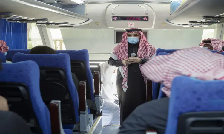 A man stood in front of the Sudanese Liberation Front with chairs, wearing a Saudi ghutra with a shemagh, an aqal and a Saudi bisht, as well as a medical mask. He was holding a microphone in his hand and speaking solemnly to the seated thermos, as part of the Umrah program organized by Qassim University accompanying the martyrs.