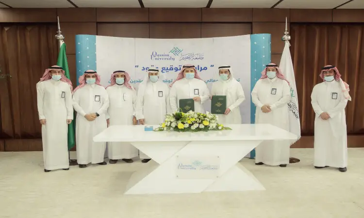 يجتمع عدد من الرجال مرتدين الغترة أو الشماغ السعودي مع العقال، ويرتدون البشت السعودي حول طاولة بيضاء تحمل مستندات رجالي، حيث يلتقطون صورة تذكارية خلال مراسم توقيع عقد التشغيل والصيانة في جامعة القصيم. للحفاظ على الخلفية وعلامات الإشارات والرموز الرسمية للفعاليات.