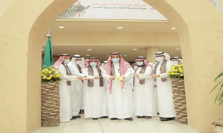 مجموعة من الرجال الحاضرين الغترة أو الشماغ السعودي مع العقال والبشتاء السعودي يشاركون في مراسم قص الشريط تحت قوسٍ في إحدى المواجهات في القصيم، حيث تتوزع الزهور على الداعمين وتتوسط المكان مكتوبة باللغة العربية. لحضور هذه الاحتفالية بمناسبة يوم الذكرى السنوية لتاريخ الدولة السعودية.