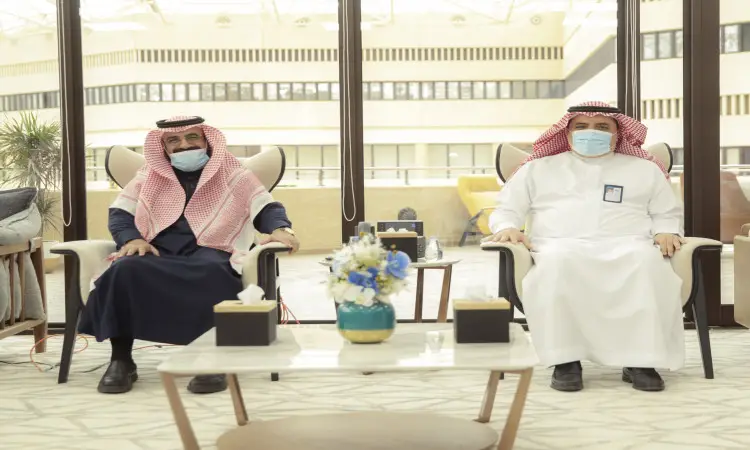 رجلان يرتديان الغترة أو الشماغ والعقال والبشت السعودي، ملتزمون بارتداء الكمامات، في مكان مغلق داخل جامعة القصيم. ويتوسطهما طاولة صغيرة عليها أكياس الزهور وصناديق المناديل، مما يؤكد صحة الجودة. وتطلّع خلفهم نوافذ كبيرة لتبني مبنى حديثاً يعكس العصر الرقمي للجامعة.