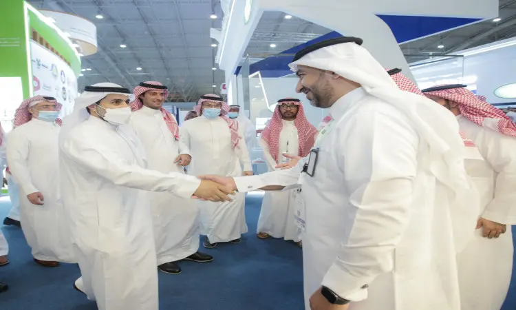 A number of men wearing the Saudi Ghutra or Shemagh, along with the Saudi Aqal and Bisht, gather in a house in Riyadh, some wearing masks. They exchange greetings and shake hands in the center of the room, keeping behind them the pavilions and banners of the Middle East Poultry Exhibition.