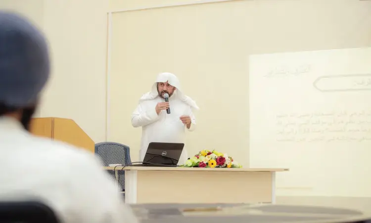 A man wearing a Saudi ghutra, a Saudi qalq and a Saudi bisht, speaks through a megaphone to the audience next to a laptop and a bouquet of flowers. Behind him is a screen displaying a text in Arabic on the topic of intellectual fortification, in the audience hall in Qassim.