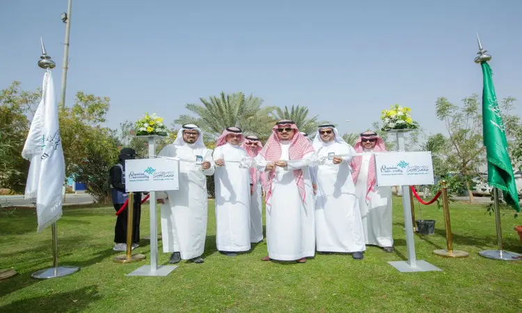 A group of men dressed in Saudi Arabian ghutra or shemagh with an aqal and Saudi bisht, connect outdoors on green lawns during a tree-planting ceremony at Qassim University. They show a smiling ribbon as they begin the inauguration, surrounded by two flag poles adorned with joins and no flowers on the links, while the works and a clear sky look on.