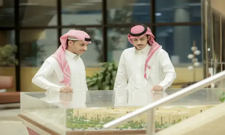 Two men dressed in Saudi Arabian ghutra, headband and bisht contemplate an architectural model in a modern unit in Qassim, where new windows and plants are visible in the background, a scene that emphasizes the refreshments for the start of the new university.