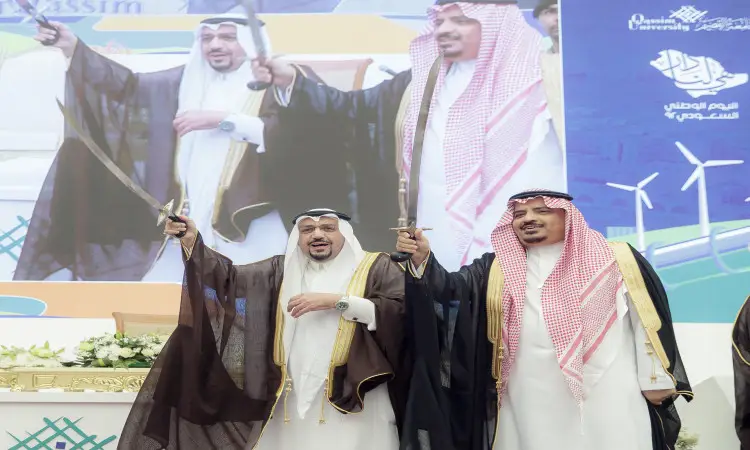 خلال جامعة القصيم، عصران يرتدي رجلان الغترة أو الشماغ السعودي مع العقال والبشت السعودي السيوف عالياً بابتسامة فخر وعتزاز. في الهواء الطلق، صورهم الخلفية على شاشة كبيرة برسومات توربينات الرياح ونص باللغة العربية يبرز شعار اليوم الوطني 92.
