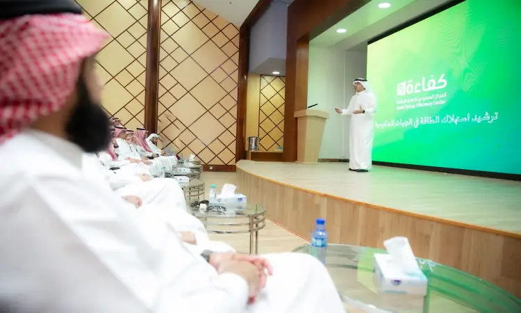 Cat Man wearing the Saudi Arabian Ghutra, Eqal and Bisht on stage, as an adult, during a workshop for government entities at Qassim University. Behind him, a large green screen displayed text in Arabic and part of the phrase "Knowledge Learning Center." They took their seats, facing the stage with concentration and attention.