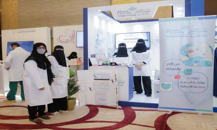 ألت عدد من الأشخاص بالزي الطبي في جناح كلية التمريض بمعرض مقام في جامعة القصيم. تتواجد سيدتان ترتديان ملابس بيضاء وتضعان كمامات طبية في حماية الجناح، بينما تلبس خلف الستار ثلاثة موظفين وتلبس الغترة أو الشماغ والعقال، ويرتدي التضامن البشت الرسمي السعودي. تتزين المساحة بمواد تعريفية ولافتات تسلط الضوء على الكونجرس السعودي للفرد، مما يعكس المعلومات الرسمية للطابعة والفخامة التي تحلى بها الجامعة في تنظيم الفعاليات.