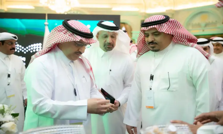 اتصل بمجموعة من الرجال مرتدين الغترة أو الشماغ مع العقال والبشت السعودي في مكان داخلي خلال مؤتمر الشراكات والعمل. يقوم أحد بعرض شاشة عرض على هاتفه المحمول لاثنين من الاثنين، بينما يتابع عدد من المشاهدين بالقرب من جناح جامعة القصيم.