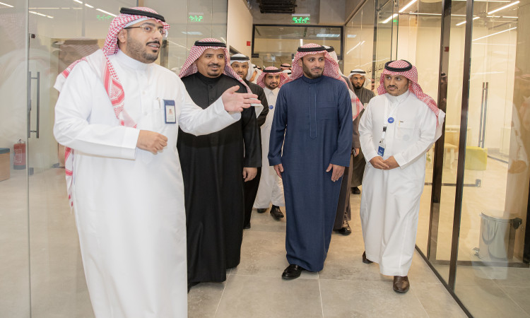 يسير مجموعة من الرجال تحت الغترة أو الشماغ والعقال والبشت السعودي داخل مبنى إداري حديث في جامعة القصيم يتميز بجدرانهزجاجات وإضائته الساطعة، ويتبادلون النقاش حول ريادة الأعمال الرقمية، بينما يتقدمهم أحدهم ملوحًا بيده أثناء حديثه الرسمي مع المجموعة.