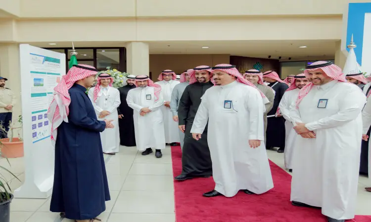 أت مجموعة من الرجال مرتدين الغترة السعودية والشماع والعقال والبشت السعودي على بساط أحمر، يستمعون إلى مقدم المشاهير يرتدي بشتاً أزرق وشماغاً سعودياً ضمن ملتقى طلبة الدراسات العليا، وذلك في أروقة مبنى حديث في القصيم تزينه ملصقات تعريفية وإرشادية.