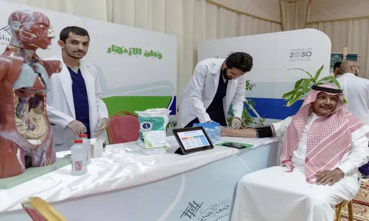 Two men dressed in white coats, wearing a Saudi shemagh, check the blood pressure of a visitor at a health corner within the Buraydah Spring Festival 44. The corner features health options and modern medical supplies, along with a tablet that displays a health education information form. The official 2030 slogans and Arabic texts benefit from the participation of a student club from Qassim University, where participants are also seen wearing the Saudi bisht in a scene that replicates the national identity and official stamp of the occasion.