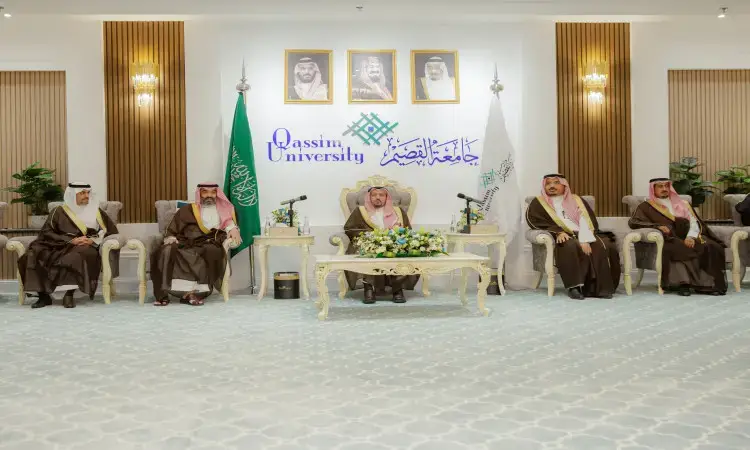 An official meeting at Qassim University, attended by His Highness the Emir of Qassim and four other Saudis, wearing the Saudi Shemagh, Ghutrah, Eqal and Bisht, sitting on plush chairs. Saudi Arabia's flags and leadership portraits shine in the background, along with the Qassim University logo in part.