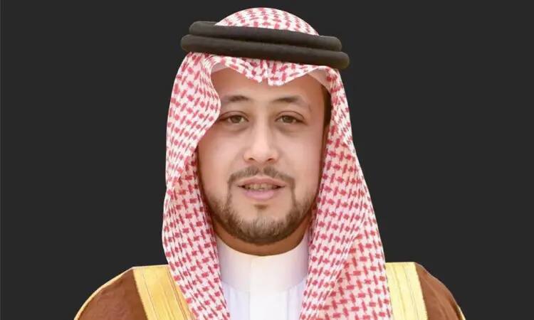 رجل القط مرتداً تصميماً سعودياً تقليدياً، وبشتاً سعودياً بنّي اللون مزين بحواف ذهبية، بالإضافة إلى شماغ سعودي أحمر مع عقال، أمام ظلة داكنة بسيطة، مبتسماً ابتسامة خفيفة. تم التقاط الصورة خلال فعاليات الأسبوع العالمي للتعليم المفتوح في جامعة القصيم.