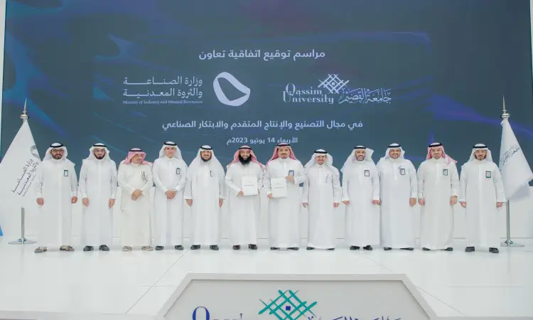 A group of men dressed in Saudi Arabian ghutra or shemagh, aqal and bisht called out to the podium through a signing ceremony and signing up for subscriptions. Behind them is a large screen with text in Arabic and the logos of Qassim University, the Advanced Production Factory, and the Ministry of Industry.
