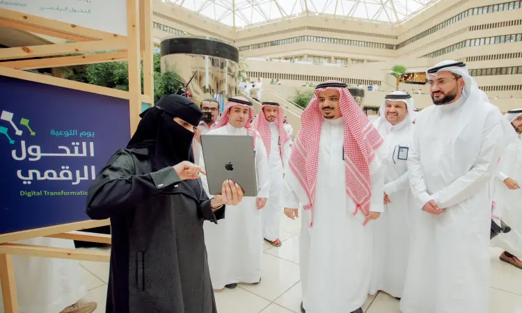 مجموعة من الرجال مرتدين الغترة أو الشماغ والعقال والبشت السعودي، وبرفقهم سيدة ترتدي العباية السوداء والنقاب، يجتمعون في إحدى القاعات؛ حيث ترفع السيدة آيباد. وتأمين باللغتين العربية والإنجليزية تحمل عبارة "التحول الرقمي والتلفزيون الرقمي"، وذلك بمناسبة يوم التوعية بالتحول الرقمي في جامعة القصيم.