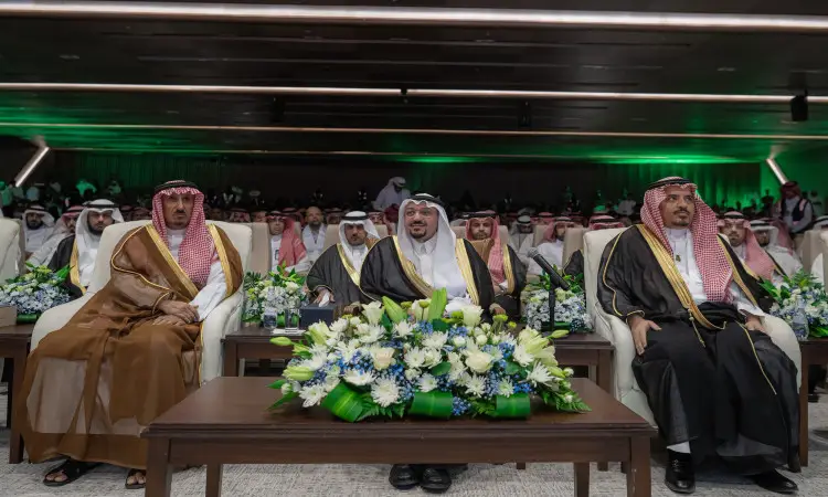 A group of men wearing the Saudi ghutra or shemagh with the Saudi aqal and bisht, led by His Highness the Emir of the region, in the front row of one of the official National Day events organized by Qassim University. The presence of the zahriyah gives the tables a distinctive formal look, as the audience in the hall behind them is crowded with a wide participation of university staff and invitees.