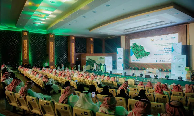 يجلسون جمع من الحضور مرتدين الغتر والشماعات والعُقُل والبشوت السعودية التقليدية في قاعة حديثة بإضاءة خضراء في جامعة القصيم، يتابعون أحداً على المنصة خلال فعاليات الملتقى الثاني "التكاتف"، بينما تعرض على شاشة الشاشة المملكة العربية السعودية ونص باللغة العربية.