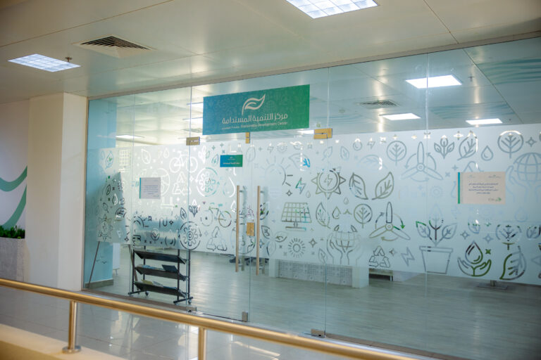 A glass-walled office decorated with eco-themed graphics and Arabic phrases, including the words "Center for Sustainable Development" written on the glass, a file rack inside the office, and a bright ceiling light that illuminates the space.