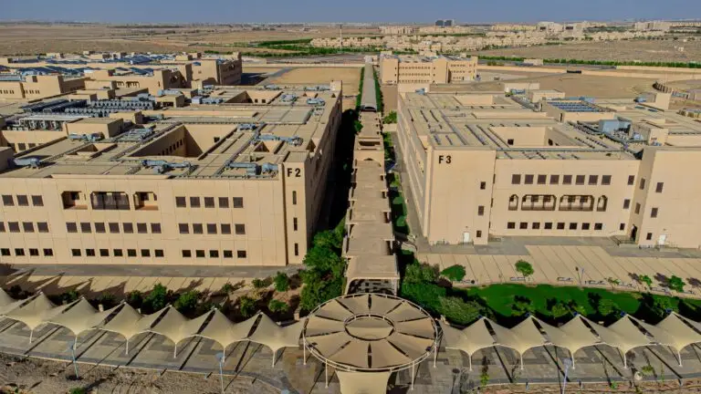 Aerial photos of two large beige-colored university buildings at Al-Qassim University, the College of Designs (and 2) and Building (and 3), separated by a corridor with geometrically designed umbrellas, and surrounded by green spaces in a desert environment.
