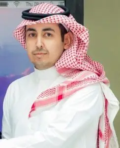 يرتدي رجل يرتدي زيًا أبيض تقليديًا مع شماغ سعودي أحمر وأبيض داخل أحد المباني، مسجون في الكاميرا بتعبير حيادي كما لو كان يلتقط صورة لملف أعضاء هيئة التدريس في جامعة القصيم.
