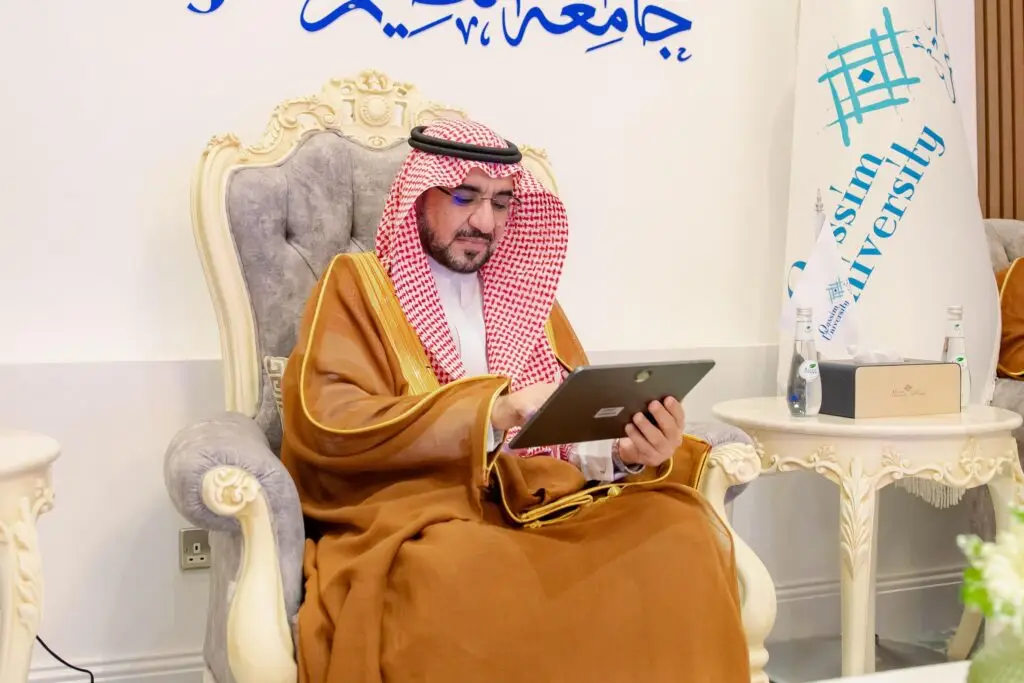 The man, dressed in traditional Saudi dress with a Saudi shemagh, sits in a plush chair, looking at a tablet for automatic masuda extraction. He takes a banner that reads "Qassim University," with more information in Arabic emblazoned on the wall.