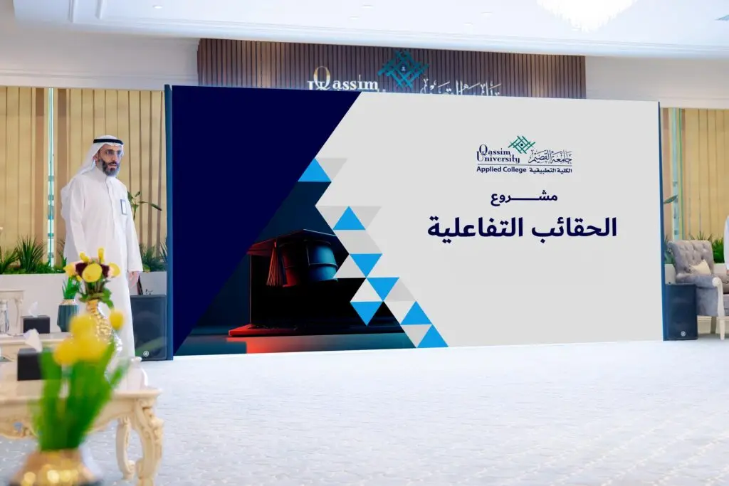 A man wearing a Saudi uniform with a Saudi shemagh or ghutra and a large screen showing text in Arabic and the logo of the Applied College, in the modern decorative hall with decorative plants and flowers.