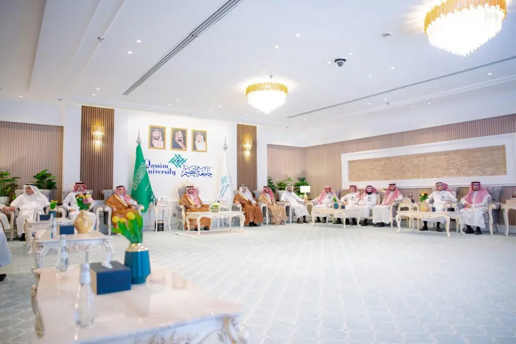 A group of men wearing the Saudi Shemagh or Saudi Ghutra meet in a hall in addition to the Applied Kalabiya in Qassim, with the flags of the Kingdom of Saudi Arabia, the head of the leadership and the university logo adorning the hall behind them. The group is discussing interactive bags at home.
