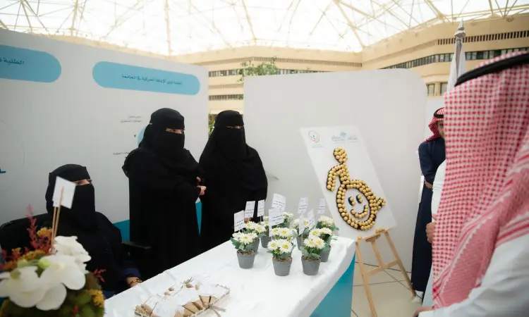 ثلاث سيدات يرتدين العباءة والنقاب يعتن خلف عرض لمنسقات وزهور، حيث يشاركن مشروعهن ضمن فعاليات التقرير العاشر. ويرتدي الرجل زياً سعودياً تقليدياً مع شماغ سعودي، متابعاً للعرض، للاستهلاك في إحدى وجهات جامعة مباني.