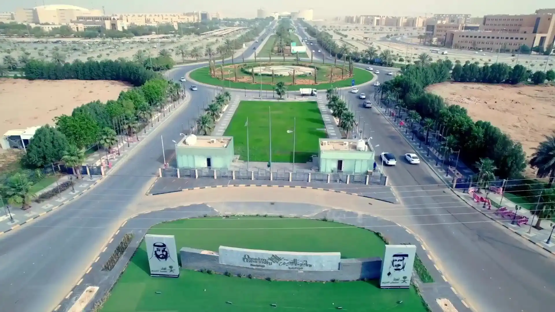 An aerial view of a landscaped garden within the university campus, which includes extensive greenery, trees and merchants, surrounded by internal roads with limited vehicular traffic, a central roundabout in the center of the image, and Qassim University buildings in the background.