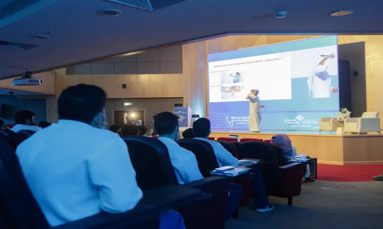 يُسيطر على الحضور في قاعة المحاضرات، وهو يعتمد على شاشة عرض كبيرة لقطاعات العرض، بالإضافة إلى صورة التركيز لضبط النفس. يتابع خطون باهتمام تقديم تقرير الهدف الرابع، وذلك ضمن فعاليات جامعة القصيم. ويلاحظ أن بعض الحضور بارتداء الشماغ السعودي أو الغترة السعودية بما في ذلك ما يليق بالطابعة الرسمية للمناسبة.
