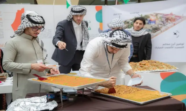 يقوم عدد من الرجال الذين تأخروا الشماغ السعودي أو الغترة السعودية بتقديم وتشكيل صوانٍ كبير من الأطباق المتنوعة ذات الألوان الزاهية من خلال الفعالية الثقافية، وذلك وذلك في ظل وجود ملصقات تعريفية وتقارير حول الهدف الرابع في الخلفية.