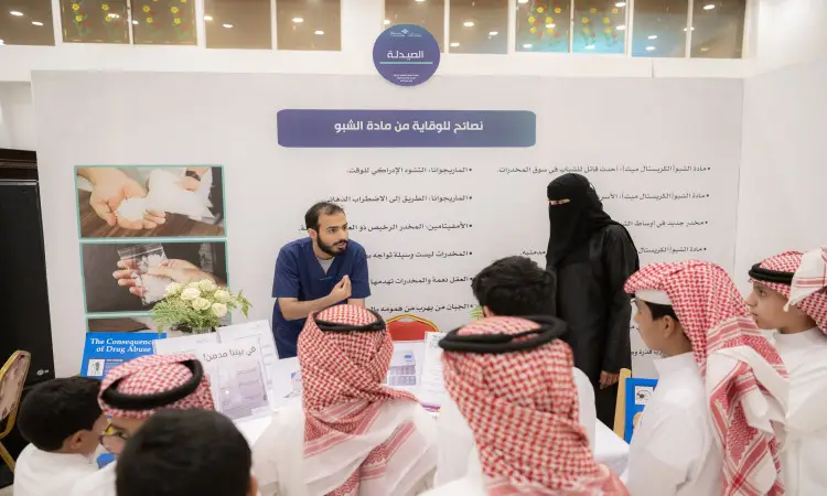 رجل يتحدث وامرأة إلى مجموعة من الناشطين الثياب الوطنية والشماغ أو الغترة السعودية في ركن توعوي ضمن فعاليات القصيم للتوعية بأضرار المخدرات، النبيذ الركن ملصقات وطويات تعليمية بالإضافة إلى تقرير يدعم الهدف الثالث.