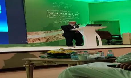 احتاج الرجل إلى كلمة من على منصة في قاعة ذات خلفية خضراء ونصًا باللغة العربية وتقرير الهدف الثالث عشر، وذلك خلال ندوة حول الزراعة في جامعة القصيم. تقدم في المقدمة الجداول عليها مشروبات ووجبات خفيفة.