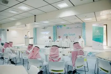 A number of men wearing Saudi Arabian shemaghs or ghattaras gather around tables and on stage in a modern conference room for a community event or panel discussion dedicated to the Goal 12 report theme. Posters, backgrounds, text and logos in Arabic are recommended.