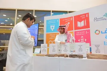 Two men dressed in traditional Saudi attire, in the form of a shemagh or Saudi ghutra, at a booth with labeled displays, where they present a report on the 12th Goal. Behind them are awareness posters and a plaque bearing the name of Qassim University. He interprets the content of the report while others follow what is displayed on the table.
