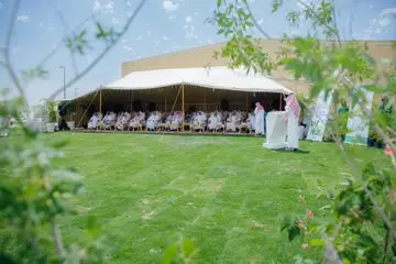 A number of people wearing the Saudi shemagh or Saudi ghutra are gathered under a large tent on a sunny day, while lush greenery and plants are in the fray. Apparently, they have announced the neighboring public speaking platform, as part of an all-day event at Qassim University.