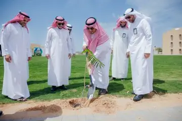 All five men are dressed in traditional Saudi attire, and the group is wearing the Saudi shemagh or Saudi ghutra, on a green space on campus. He is planting a small sapling with a shovel, while following the correct method intently, in a scene that reflects the 12th goal. Searching for a university mane and the clear blue sky.