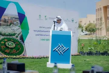 A man dressed in traditional Saudi dress with a Saudi shemagh stands behind a platform to face an open-air speech, behind him is a board with text in Arabic and the word "report" as well as a site plan. To prepare simple information about some plants and buildings on a sunny day, as part of a children's event in Qassim.
