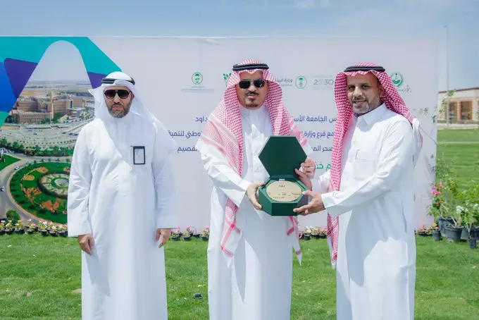 Three men in authentic Saudi uniforms, the man in the center is handing a green honorary shield to the man on his right, in celebration of the 12th goal. In the background are some flowers.