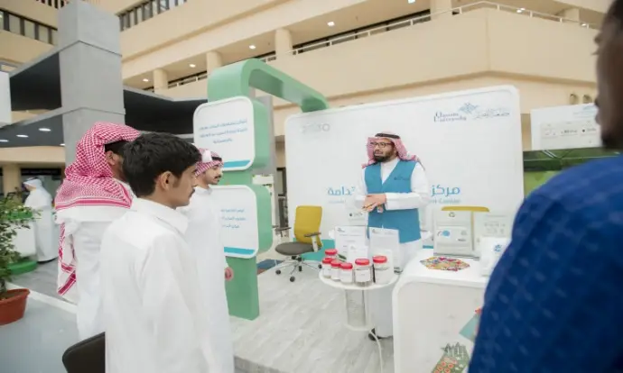 A group of men in Saudi Arabian shemaghs and robes gather to listen to the presenter in an informational booth inside a modern building. Informational posters for beginners in Arabic are a highlight of the show, with templates, cases, brochures, and a report prepared for the show.