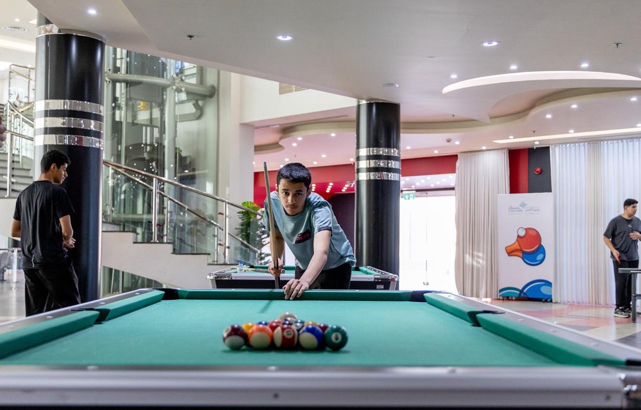 A young man prepares to shoot a shot in the game of billiards inside the summer club of Qassim University, where he directs the cue of the set of balls carefully arranged on the table in a modern and lighted area that witnesses the presence of many people and the organization of multiple events in the background. It should be noted that the participants - according to university customs - are obliged to wear the Saudi shemagh or Saudi ghutra while participating in these events.