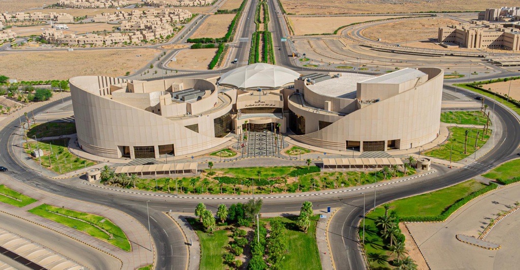 An aerial view of the King Abdulaziz Center, one of the most prominent modern landmarks at Qassim University. It is characterized by a unique architectural design that includes curves and a central dome that surrounds the building with neatly organized streets, lush green spaces and carefully coordinated gardens, with desert areas interspersed with other facilities belonging to the university in the background.
