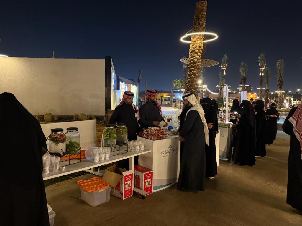 A health awareness event at Qassim University in an outdoor plaza, distributing drinks and food under a lighted palm tree.