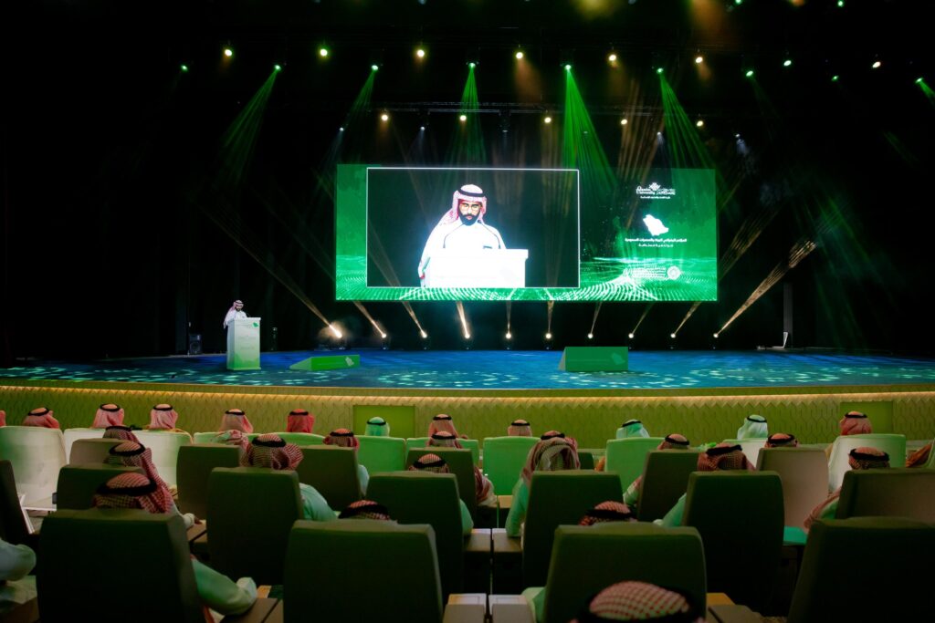 إلقاء كلمة في المؤتمر الجغرافي الجغرافي بجامعة القصيم بحضور أكاديمي في قاعة مضاءة بالأخضر.